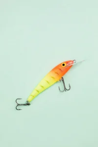 Bright orange and yellow artificial fishing lure with treble hooks on a light green background.