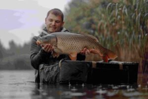  A smiling angler holding a large carp fish near the water, surrounded by greenery